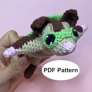 Puede incluir: Juguete de amigurumi tejido a crochet de una ardilla voladora con gafas verdes. El juguete es marrón y beige con una nariz rosa. El texto "PDF Pattern" es visible en la imagen.