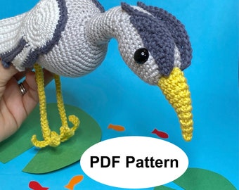 May include: Crochet pattern for a gray and yellow heron amigurumi toy. The pattern is available as a PDF download. The toy is sitting on a green lily pad with red and orange fish.
