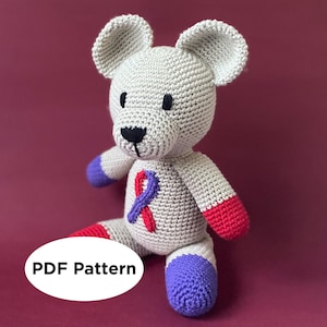 May include: A crocheted gray teddy bear with a purple and red ribbon on its chest. The bear has a black nose and eyes. The bear's feet are purple and red.