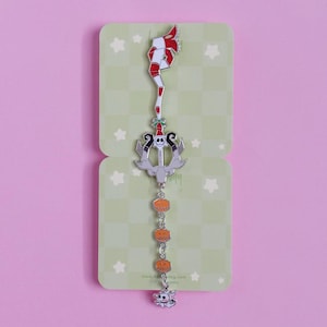 May include: A decorative metal bookmark featuring a whimsical design. The top has a red and white striped element, followed by a silver trident with a skull, and a chain of orange pumpkin charms. The bottom has a skull charm. The bookmark is on a light green card.