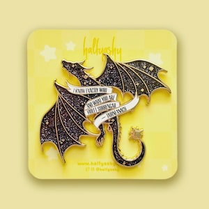 May include: A black glittery dragon enamel pin with gold metal outlines. A white banner across the dragon features the black text: I KNOW EXACTLY WHO AND WHAT YOU ARE VIOLET SORRENGAIL - TAIRNEANACH. The pin is attached to a yellow backing card.