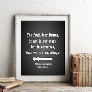 Puede incluir: Una impresión en blanco y negro enmarcada con una cita de Julio César de William Shakespeare: "La culpa, querido Bruto, no está en nuestras estrellas, sino en nosotros mismos, que somos subalternos." La impresión está en un estante con libros y un reloj.