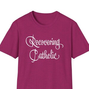 Może przedstawiać: Koszulka w kolorze magenta z napisem "recovering catholic" w białym, ozdobnym pismie.