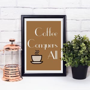 以下が含まれることがあります： 茶色の背景に白い文字で「Coffee Conquers All」と書かれた額入りのプリント。銅とガラス製のフレンチプレスと小さな鉢植えが、白いレンガの壁の前に白い表面に置かれています。