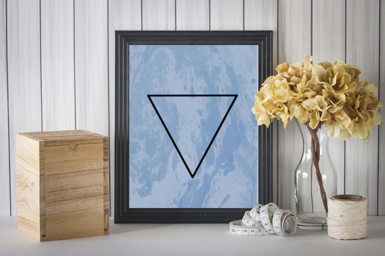 May include: A framed print with a blue and white marbled background and a black triangle in the center. The frame is black and the print is hanging on a white wood wall.