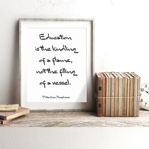 May include: A framed print with a white background and black text that reads "Education is the kindling of a flame, not the filling of a vessel. -Plutarchion Paraphrase". The print is sitting on a wooden shelf with a stack of books and a clock.