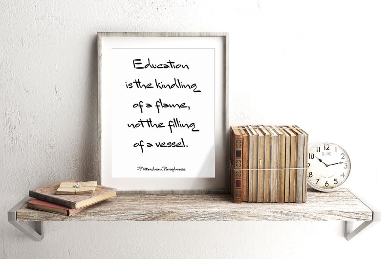 May include: A framed print with a white background and black text that reads "Education is the kindling of a flame, not the filling of a vessel. -Plutarchion Paraphrase". The print is sitting on a wooden shelf with a stack of books and a clock.