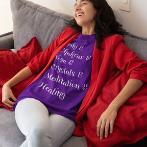 May include: A woman wearing a purple t-shirt with the text "Reiki & Chakras & Yoga & Crystals & Meditation & Healing" in white lettering. She is sitting on a gray couch with a red throw pillow.