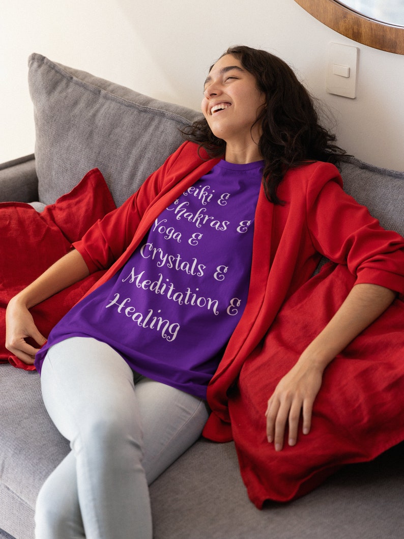 May include: A woman wearing a purple t-shirt with the text "Reiki & Chakras & Yoga & Crystals & Meditation & Healing" in white lettering. She is sitting on a gray couch with a red throw pillow.