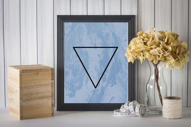 May include: A black framed print with a blue and white marbled background and a black triangle in the center. The print is hanging on a white wood wall with a wooden box and a vase of dried flowers in front of it.