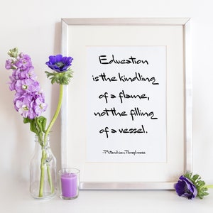 May include: A framed print with a white background and black text that reads "Education is the kindling of a flame, not the filling of a vessel. -Plutarchian Paraphrase". The print is surrounded by a silver frame and is sitting on a white surface with purple flowers in a glass vase and a purple candle.