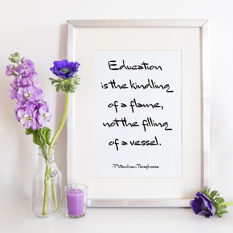 May include: A framed print with a white background and black text that reads "Education is the kindling of a flame, not the filling of a vessel. -Plutarchian Paraphrase". The print is surrounded by a silver frame and is sitting on a white surface with purple flowers in a glass vase and a purple candle.