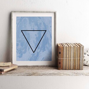 May include: A framed print with a blue and white marbled background and a black triangle in the center. The print is on a wooden shelf with a stack of books and a clock.