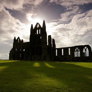 May include: Silhouette of a stone abbey against a dramatic sky. The sun shines through the abbey's arched windows and towers, casting long shadows on the green grass below. The image evokes a sense of history and grandeur.