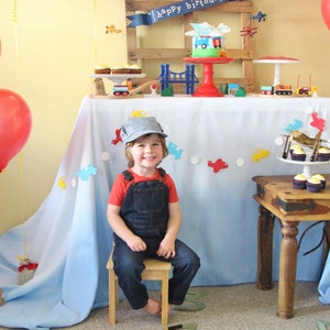 May include: A young boy wearing a blue and white engineer's cap and overalls sits on a small wooden stool in front of a birthday party table. The table is covered in a blue and white tablecloth and decorated with paper airplanes, a red and white cake, and cupcakes. The boy is smiling and looking at the camera.