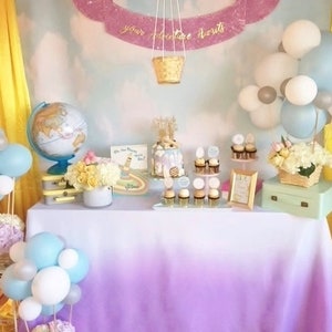 May include: A purple and white hot air balloon with a banner that says "your adventure awaits" hangs above a table decorated with a globe, flowers, and cupcakes. The table has a white and purple ombre tablecloth.