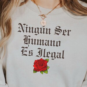 May include: A beige sweatshirt with the text "Ningun Ser Humano Es Ilegal" in black gothic font. A red rose is centered below the text.