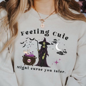 Puede incluir: Una sudadera de color beige claro con un gr&aacute;fico de una bruja y el texto "Feeling Cute Might Curse You Later." La bruja lleva un vestido morado y un sombrero negro. El gr&aacute;fico tambi&eacute;n incluye un caldero, murci&eacute;lagos, una luna creciente y estrellas.