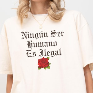 May include: A white t-shirt with the text "Ningún Ser Humano Es Ilegal" in black lettering and a red rose graphic. The shirt is being worn by a person.