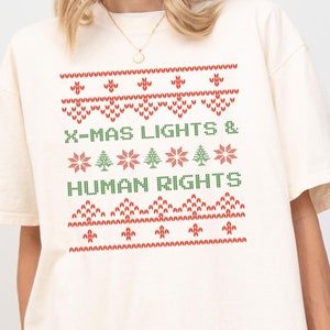 May include: A white t-shirt with a red and green Christmas-themed design. The design features a stylized Christmas tree, snowflakes, and the text "X-MAS LIGHTS & HUMAN RIGHTS".
