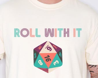 Puede incluir: Camiseta de color marfil con la frase "ROLL WITH IT" sobre un gráfico de dados multicolor. El dado muestra los números 3, 8, 16 y 19. La camiseta es de estilo casual, adecuada para el uso diario.
