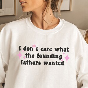 May include: White sweatshirt with the text "I don't care what the founding fathers wanted" in black letters with pink stars and a pink flower.
