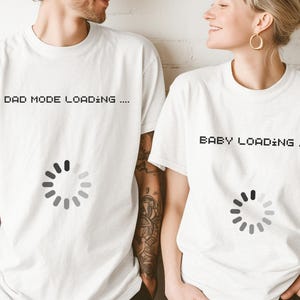 May include: Two white t-shirts with the text "DAD MODE LOADING ...." and "BABY LOADING ...." printed in black. Each shirt features a loading circle graphic. The shirts are worn by a man and a woman.