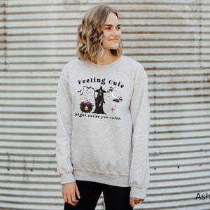 Puede incluir: Una sudadera gris con un gr&aacute;fico de una bruja y un caldero. El texto en la sudadera dice "Feeling Cute Might Curse You Later."