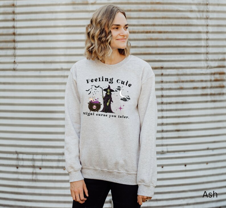 Puede incluir: Una sudadera gris con un gr&aacute;fico de una bruja y un caldero. El texto en la sudadera dice "Feeling Cute Might Curse You Later."