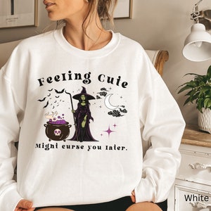 Puede incluir: Sudadera blanca con un gr&aacute;fico de una bruja revolviendo una caldera con el texto "Feeling Cute Might Curse You Later."