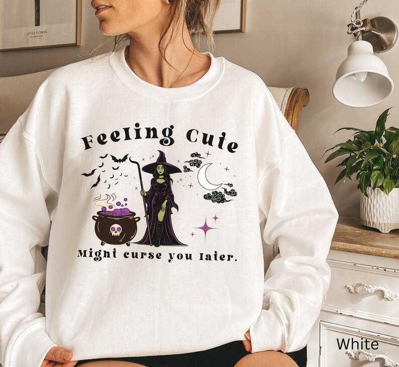 Puede incluir: Sudadera blanca con un gr&aacute;fico de una bruja revolviendo una caldera con el texto "Feeling Cute Might Curse You Later."
