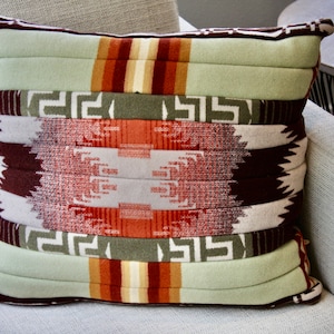 Puede incluir: Una almohada de tiro verde, marrón y blanca con un diseño geométrico. La almohada está hecha de una tela suave y mullida y tiene un brillo sutil.