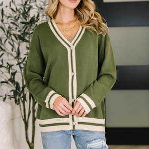 May include: An olive green V-neck cardigan featuring a cream-colored trim along the button placket, cuffs, and hem. The knit sweater has long sleeves, a button-down front with silver-toned buttons, and a ribbed texture on the edges.