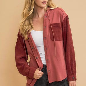 May include: A red clay-colored button-down shirt with a hood and a front pocket. The sleeves are a darker red with a textured pattern. The text "RED CLAY" is at the bottom of the image.