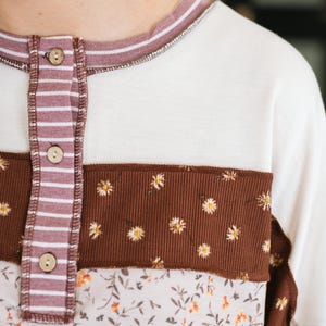 May include: A long-sleeve shirt featuring a cream-colored base with a vertical placket of mauve and white stripes. The garment includes three wooden buttons and horizontal panels of brown ribbed fabric with a small white daisy print.