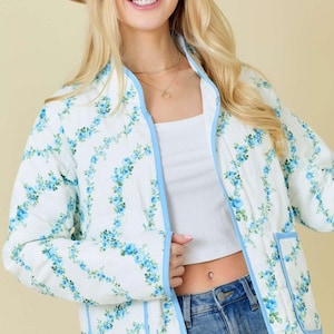 May include: A white quilted jacket with a blue floral pattern and blue trim. The jacket has a mandarin collar and a front zipper closure.