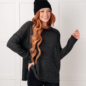 May include: A charcoal grey long-sleeved sweater with a ribbed texture, a round neckline, and side slits at the hem, paired with a solid black beanie. The sweater features visible seam detailing along the shoulders and sleeves, providing a structured look.