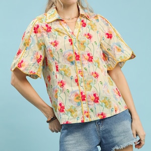 May include: Cream-colored button-down shirt with a floral pattern featuring red, pink, and yellow flowers. The shirt has short sleeves and yellow trim. Worn with denim shorts.