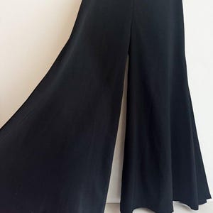 May include: Solid black wide-leg trousers featuring a high-waisted design with a center back zipper closure. The pants have a flared silhouette that extends from the waist to the hem, creating a voluminous and flowing appearance in the fabric.
