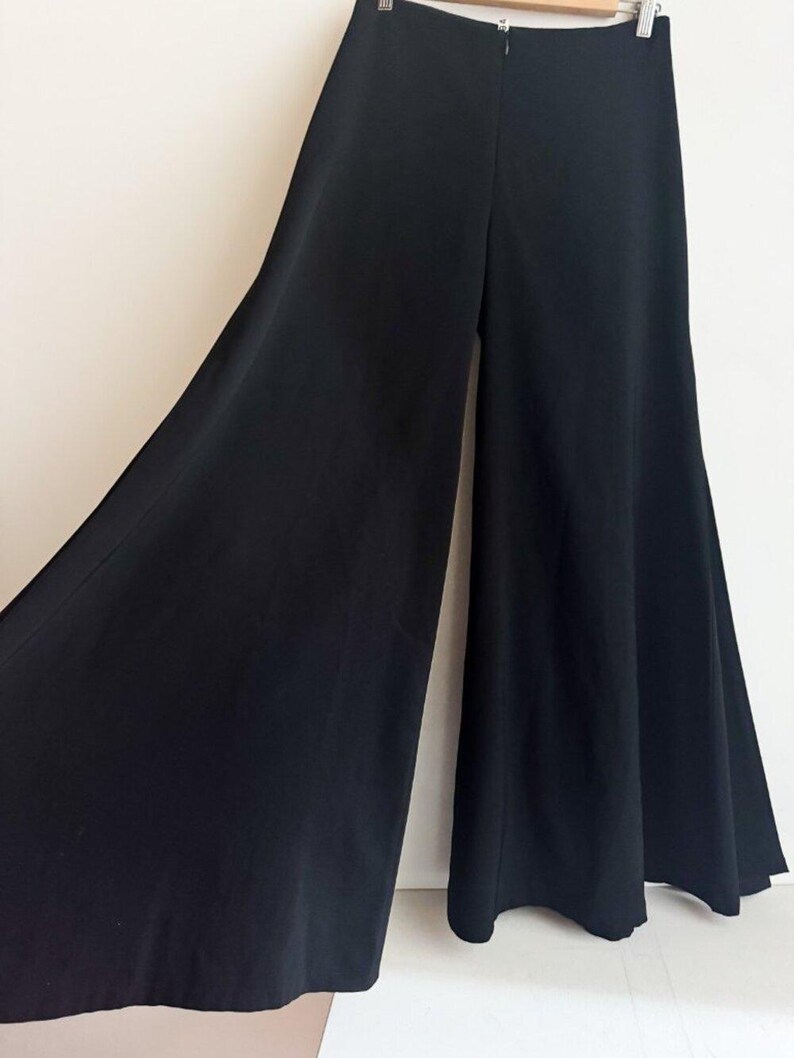 May include: Solid black wide-leg trousers featuring a high-waisted design with a center back zipper closure. The pants have a flared silhouette that extends from the waist to the hem, creating a voluminous and flowing appearance in the fabric.