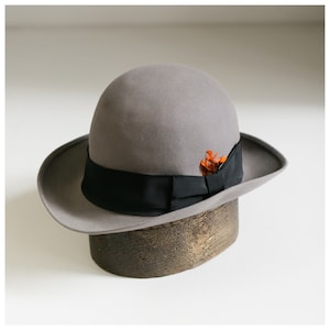 Puede incluir: Un sombrero fedora de fieltro gris con una cinta negra y un pequeño adorno de flor naranja. El sombrero está sobre un expositor de madera.