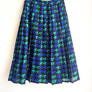 May include: A pleated midi skirt featuring a houndstooth pattern in black, teal, and blue. The skirt has a high waist and falls to mid-calf length. The fabric appears to be lightweight, suitable for warmer weather.