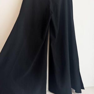 May include: A pair of solid black wide-leg trousers featuring a high-waisted design and a flowing, floor-length silhouette. The pants are constructed with a smooth, opaque fabric that drapes outward from the waist, creating a voluminous shape.