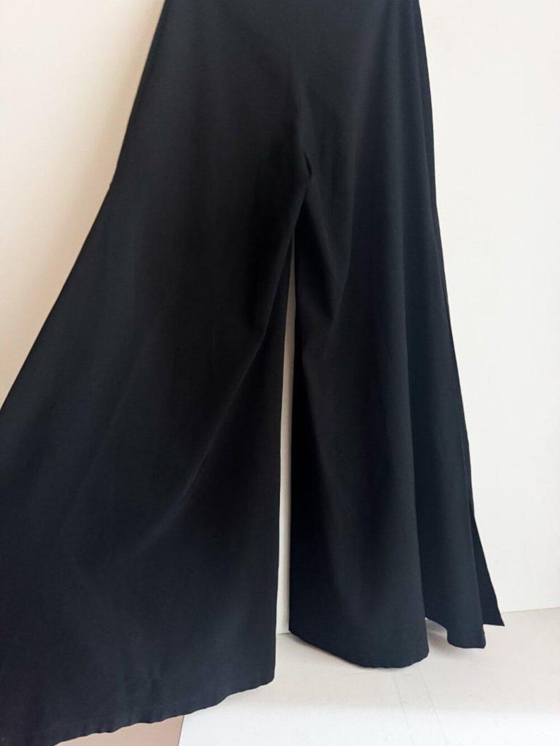 May include: A pair of solid black wide-leg trousers featuring a high-waisted design and a flowing, floor-length silhouette. The pants are constructed with a smooth, opaque fabric that drapes outward from the waist, creating a voluminous shape.