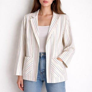 Può includere: Un blazer color crema con strisce verticali rosse, blu e viola. Il blazer ha due tasche anteriori e viene indossato aperto. La persona nell'immagine indossa jeans blu e un top bianco.