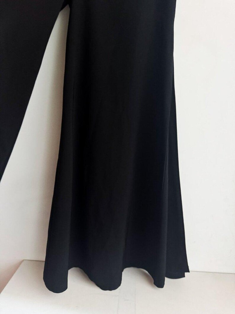 May include: A pair of solid black trousers featuring a wide-leg silhouette. The fabric is opaque and smooth, falling in soft, vertical folds from the waist down to the hem. The design is simple, focusing on the flared cut and the deep black color.
