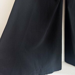 May include: A pair of solid black wide-leg trousers featuring a smooth, matte fabric texture. The garment has a loose, flowing silhouette with straight-cut legs that extend downward, showcasing a clean, unpatterned design with subtle vertical folds.