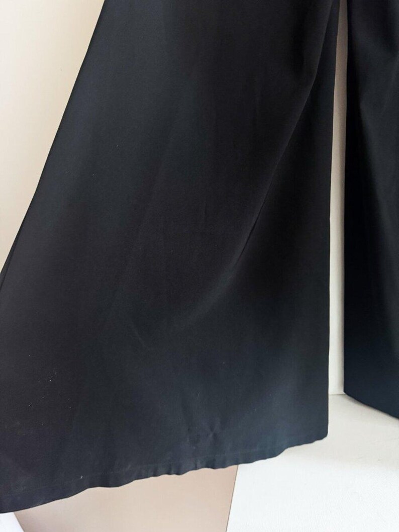 May include: A pair of solid black wide-leg trousers featuring a smooth, matte fabric texture. The garment has a loose, flowing silhouette with straight-cut legs that extend downward, showcasing a clean, unpatterned design with subtle vertical folds.