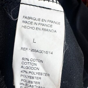 May include: A white clothing care label sewn onto a black garment. The label displays the text: FABRIQUE EN FRANCE, MADE IN FRANCE, HECHO EN FRANCIA, size L, REF: 1233AG016/14, 50% COTON, COTTON, ALGODON, 47% POLYESTER, 3% ELASTHANNE, ELASTAN, ELASTANO.