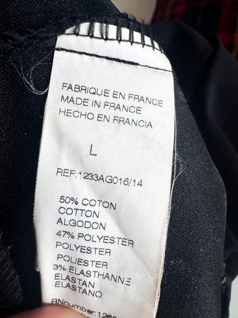 May include: A white clothing care label sewn onto a black garment. The label displays the text: FABRIQUE EN FRANCE, MADE IN FRANCE, HECHO EN FRANCIA, size L, REF: 1233AG016/14, 50% COTON, COTTON, ALGODON, 47% POLYESTER, 3% ELASTHANNE, ELASTAN, ELASTANO.